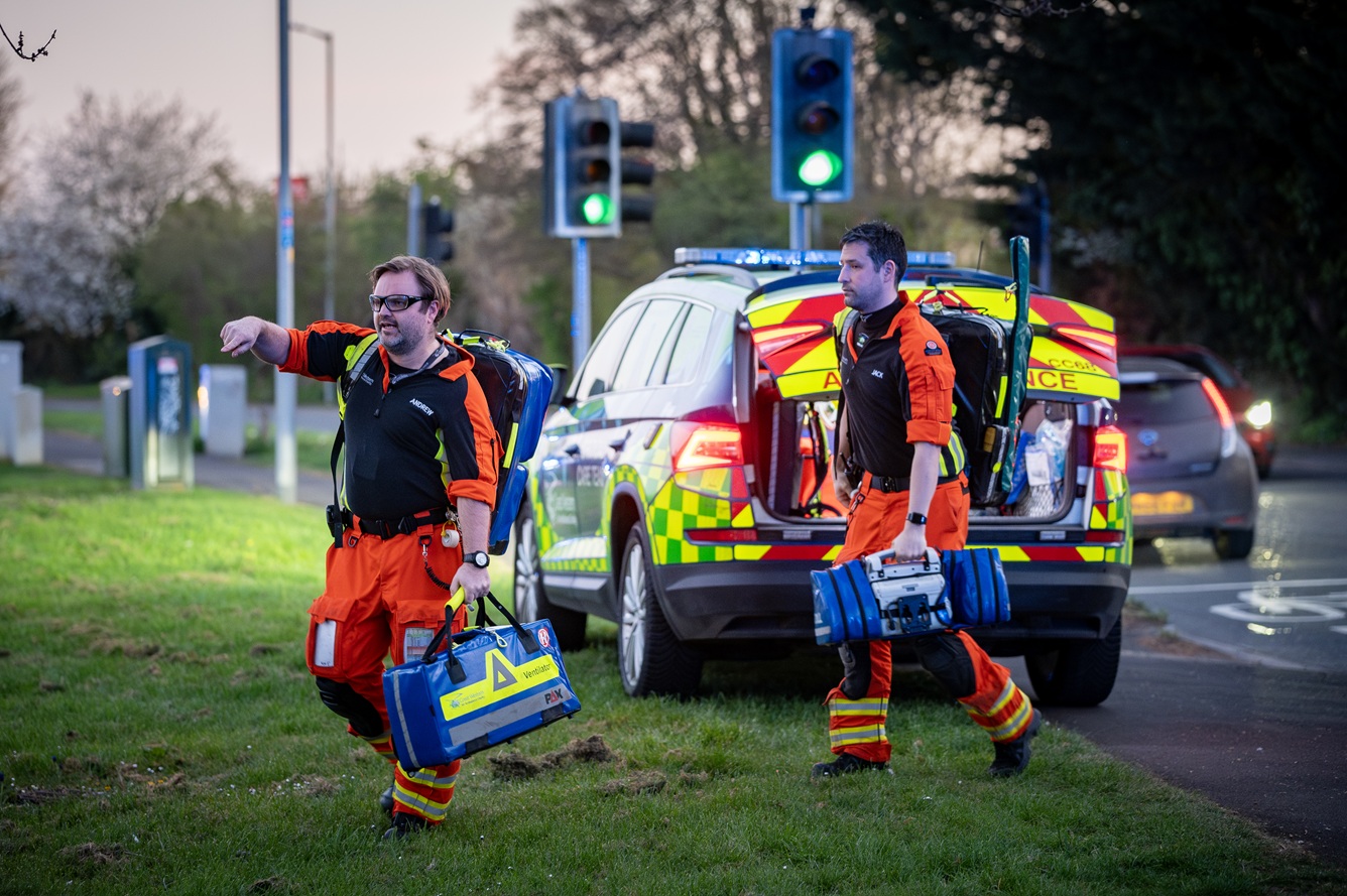 Members of the Critical Care Team arrive at the scene of an incident via their critical care car. The crew members are carrying multiple pieces of kit on their back or in their hands with the boot of the car in the process of shutting.