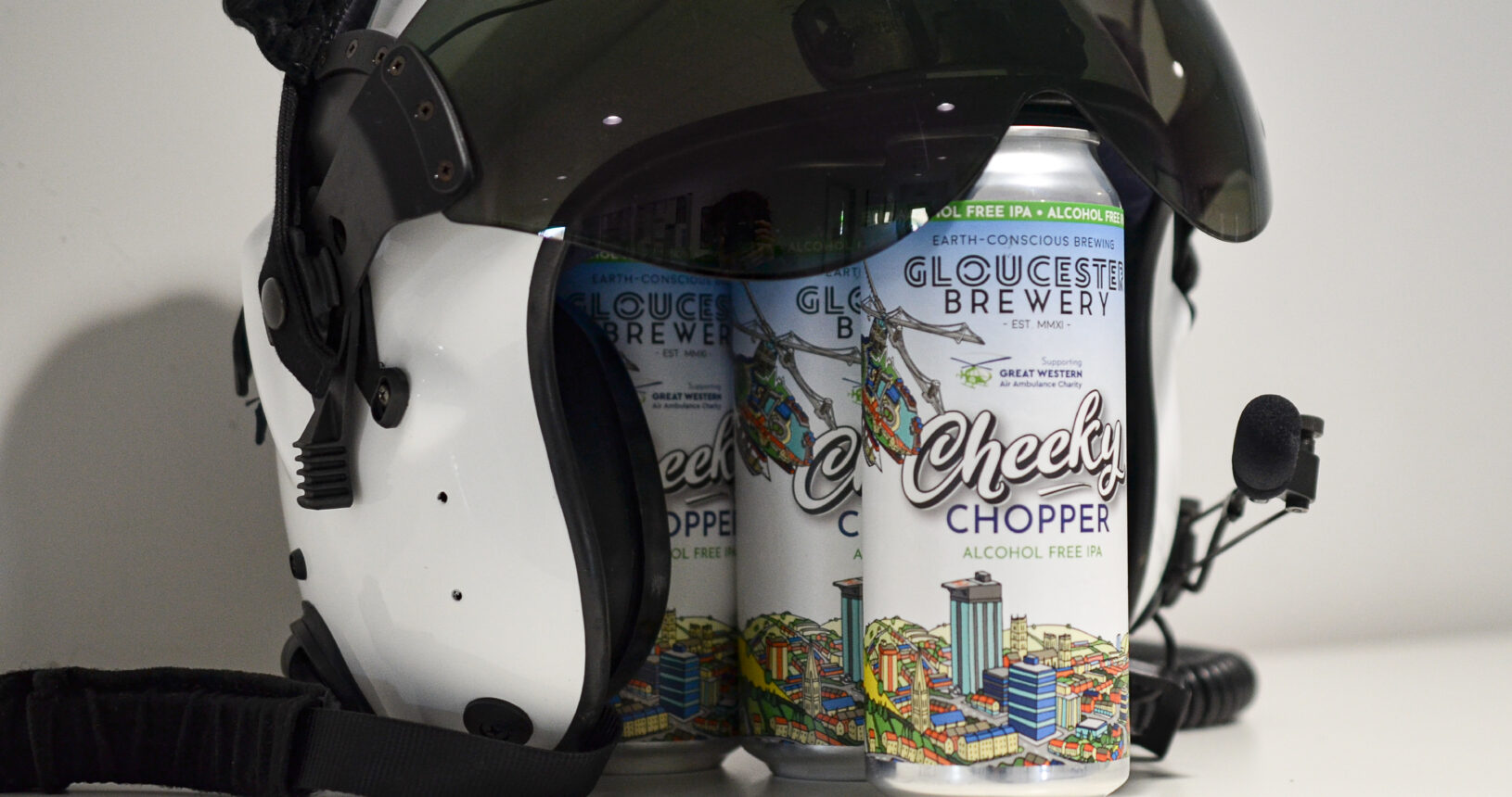 Three cans of Gloucester Brewery "Cheeky Chopper" alcohol-free IPA sitting under the raised visor of a white flight helmet, showcasing a charity partnership with Great Western Air Ambulance Charity.