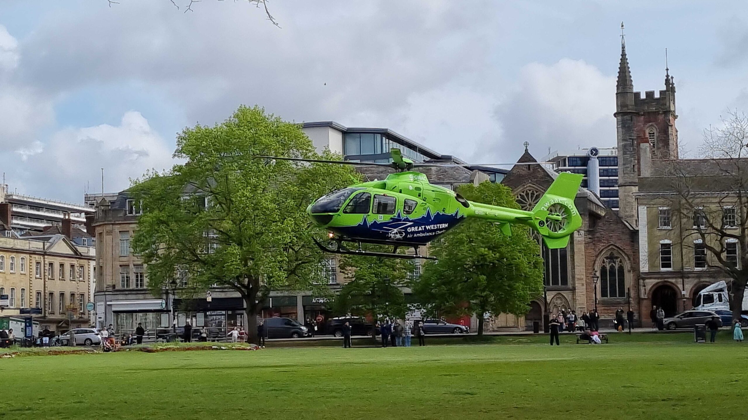 A green and blue helicopter comes into land on the field at College Green. Bristol , with buildings and trees in the background