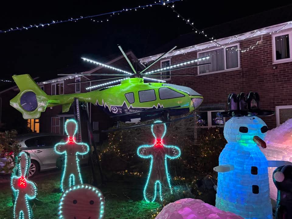 The Brookfield Walk Christmas Lights. In the image, you can see light-up gingerbread men and a snow man on the floor, with the house in the background and the main focus being the papier-mâché replica of GWAAC's green and blue helicopter, lit up in the air