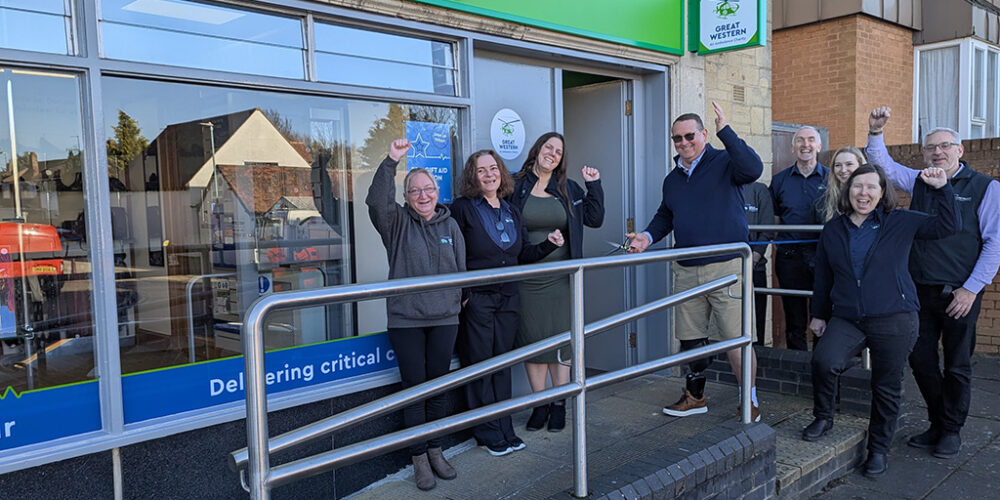 A group of people stand outside the newly opened Great Western Air Ambulance Charity shop in Brockworth, celebrating the occasion. The shop has a bright green sign with the charity's logo above the entrance. A man in sunglasses and a prosthetic leg holds a pair of scissors, having just cut the ribbon to officially open the store.