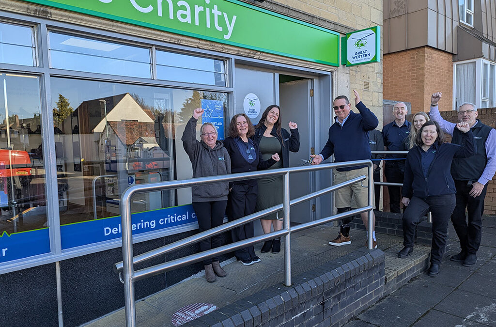 A group of people stand outside the newly opened Great Western Air Ambulance Charity shop in Brockworth, celebrating the occasion. The shop has a bright green sign with the charity's logo above the entrance. A man in sunglasses and a prosthetic leg holds a pair of scissors, having just cut the ribbon to officially open the store.