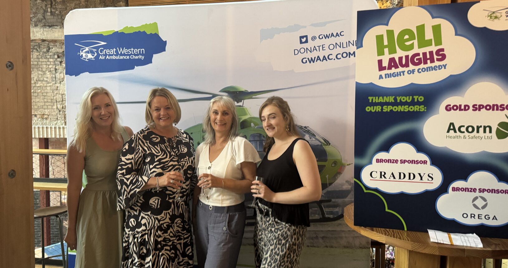 Representatives from Craddys enjoying the Heli Laughs comedy night in front of a Great Western Air Ambulance Charity banner and event sponsor board.