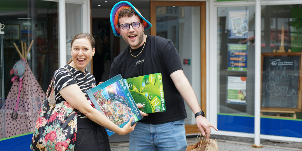 Two cheerful shoppers stand outside the Great Western Air Ambulance Charity shop in Thornbury, smiling and holding up their purchases. One person is holding a large jigsaw puzzle and a Scrabble game, while the other carries paper shopping bags and wears a colourful floppy hat. The shop entrance and signage are visible behind them.