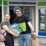 Two cheerful shoppers stand outside the Great Western Air Ambulance Charity shop in Thornbury, smiling and holding up their purchases. One person is holding a large jigsaw puzzle and a Scrabble game, while the other carries paper shopping bags and wears a colourful floppy hat. The shop entrance and signage are visible behind them.
