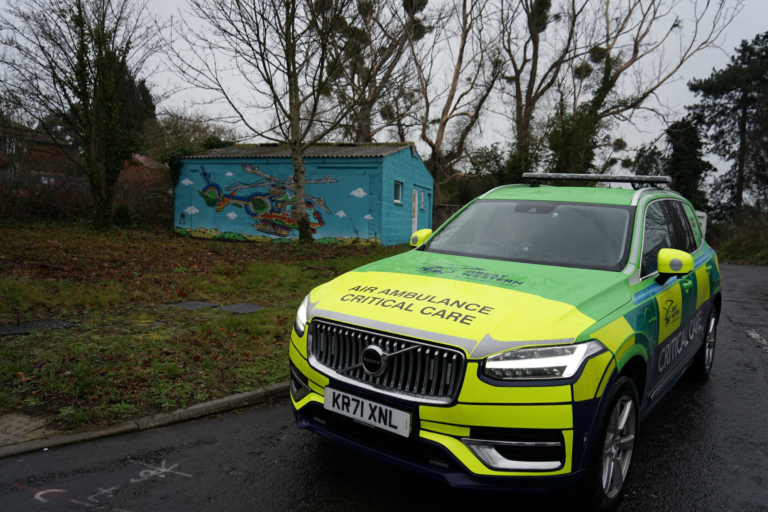 Great Western Air Ambulance Charity's new green, yellow and blue Volvo XC90 critical care car parked at the entrance to the charity's airbase in front of a  building with a helicopter mural painted on it.
