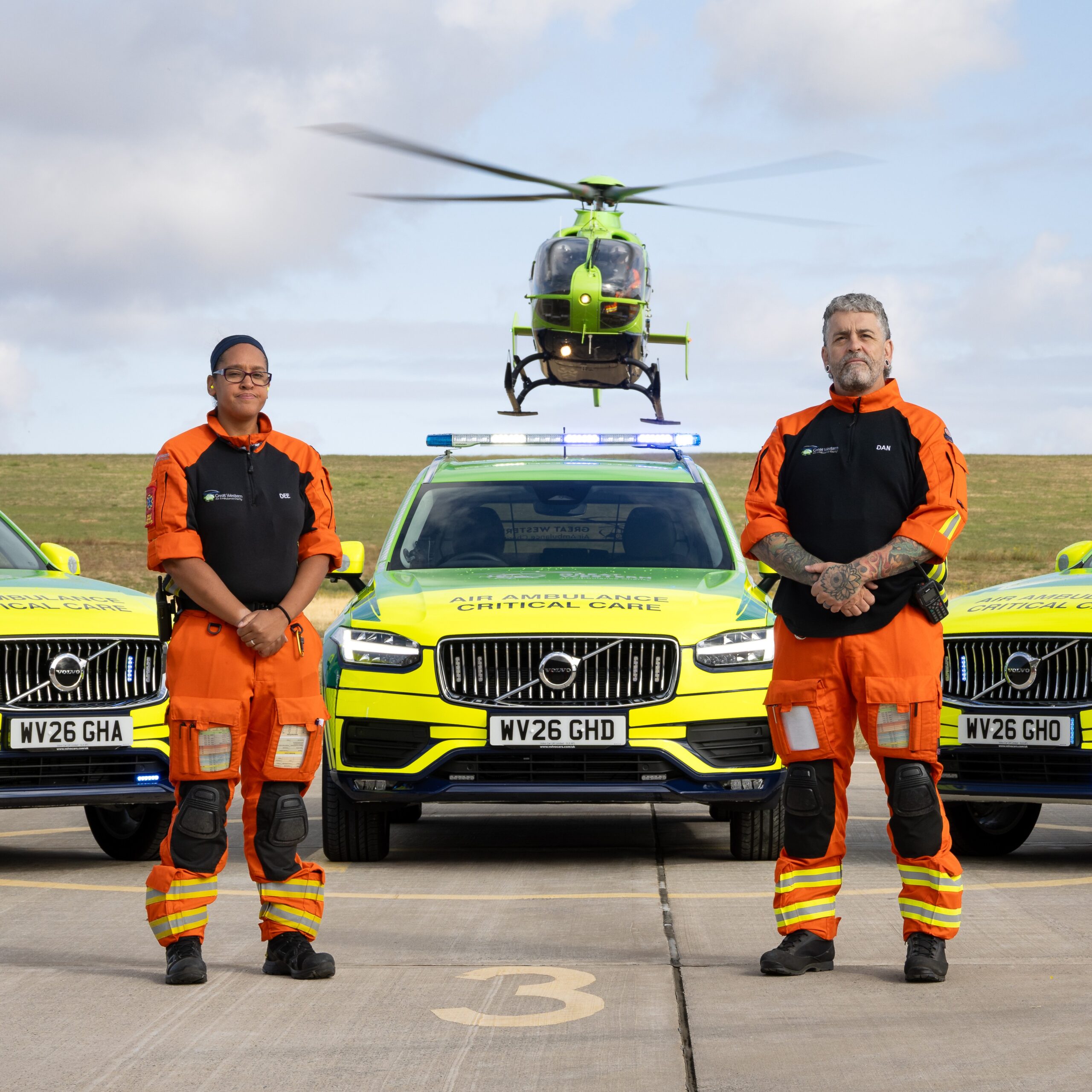 Two Great Western Air Ambulance Charity crew members standing in front of three Volvo critical care cars, with the GWAAC helicopter hovering in the sky directly above them.