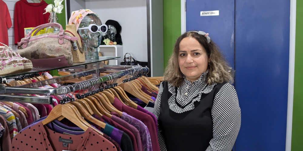 Asal, a retail volunteer at Great Western Air Ambulance Charity's Cheltenham shop, stands beside a clothing rail filled with second-hand garments, smiling at the camera. Handbags, hats and accessories are displayed on shelving behind her, with bright green walls and a blue staff door in the background.