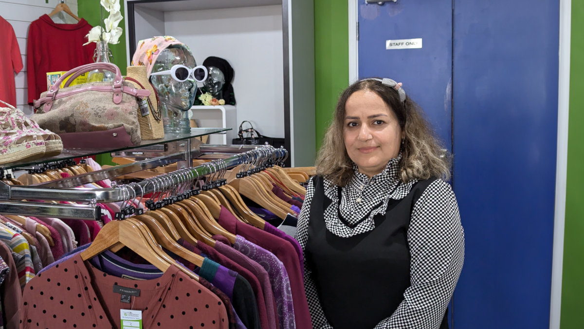 Asal, a retail volunteer at Great Western Air Ambulance Charity's Cheltenham shop, stands beside a clothing rail filled with second-hand garments, smiling at the camera. Handbags, hats and accessories are displayed on shelving behind her, with bright green walls and a blue staff door in the background.