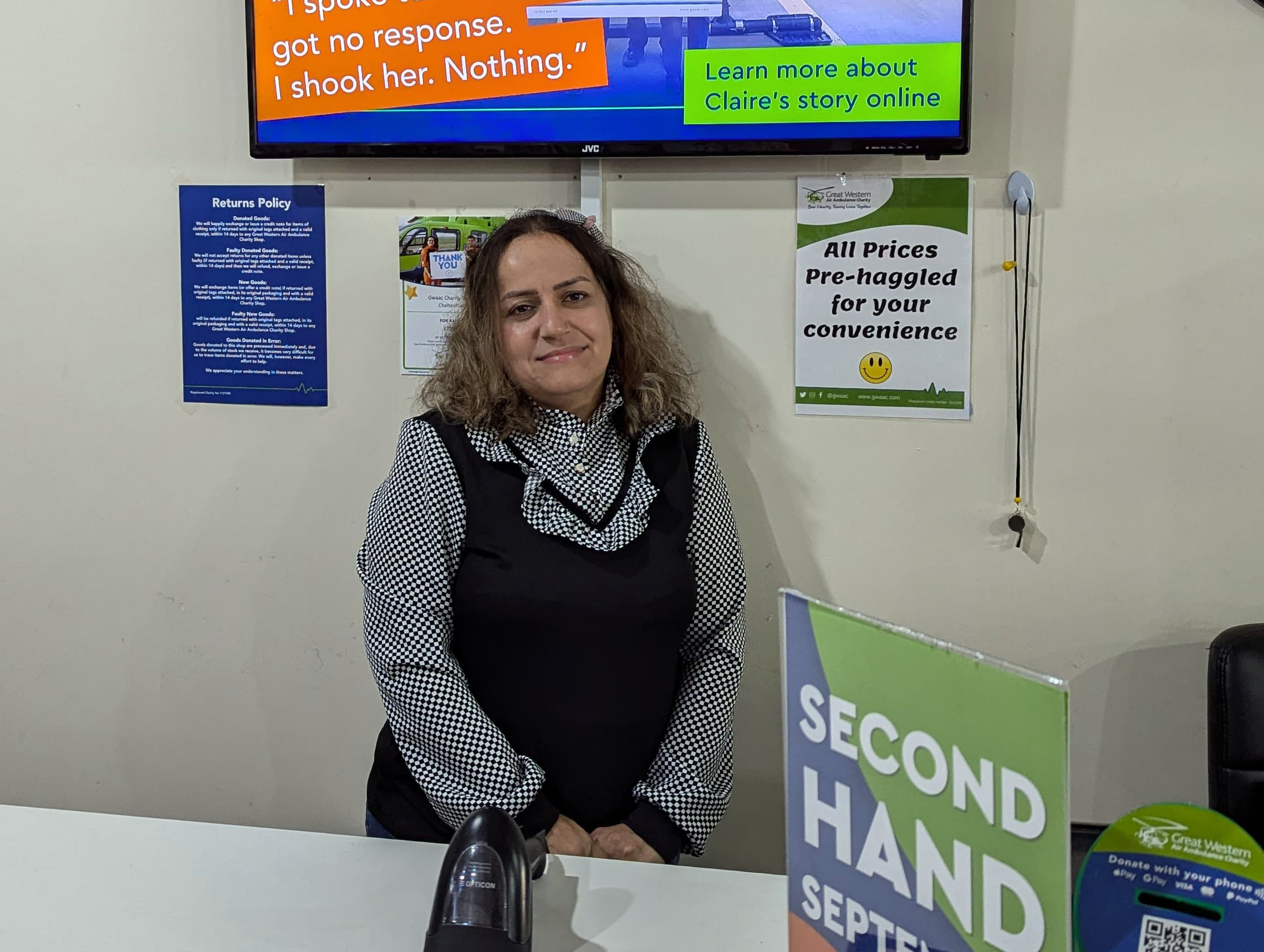 Asal, a retail volunteer at Great Western Air Ambulance Charity's Cheltenham shop, stands smiling behind the till, wearing a black-and-white patterned blouse and black pinafore. Behind her are charity posters and a TV screen, with a Second Hand September display visible in the foreground.