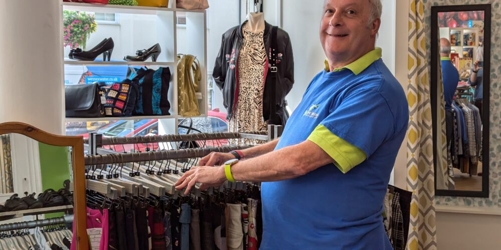 Volunteer Andy, wearing a blue and green Great Western Air Ambulance Charity t-shirt, smiles while organising a clothes rack in the Westbury-on-Trym shop. Behind him, handbags, shoes, and clothing are neatly displayed, with natural light streaming in through the window.