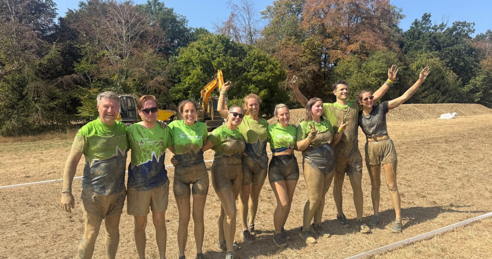 A muddy corporate team from Safran smiling and cheering in matching GWAAC t-shirts after completing a Tough Mudder challenge.