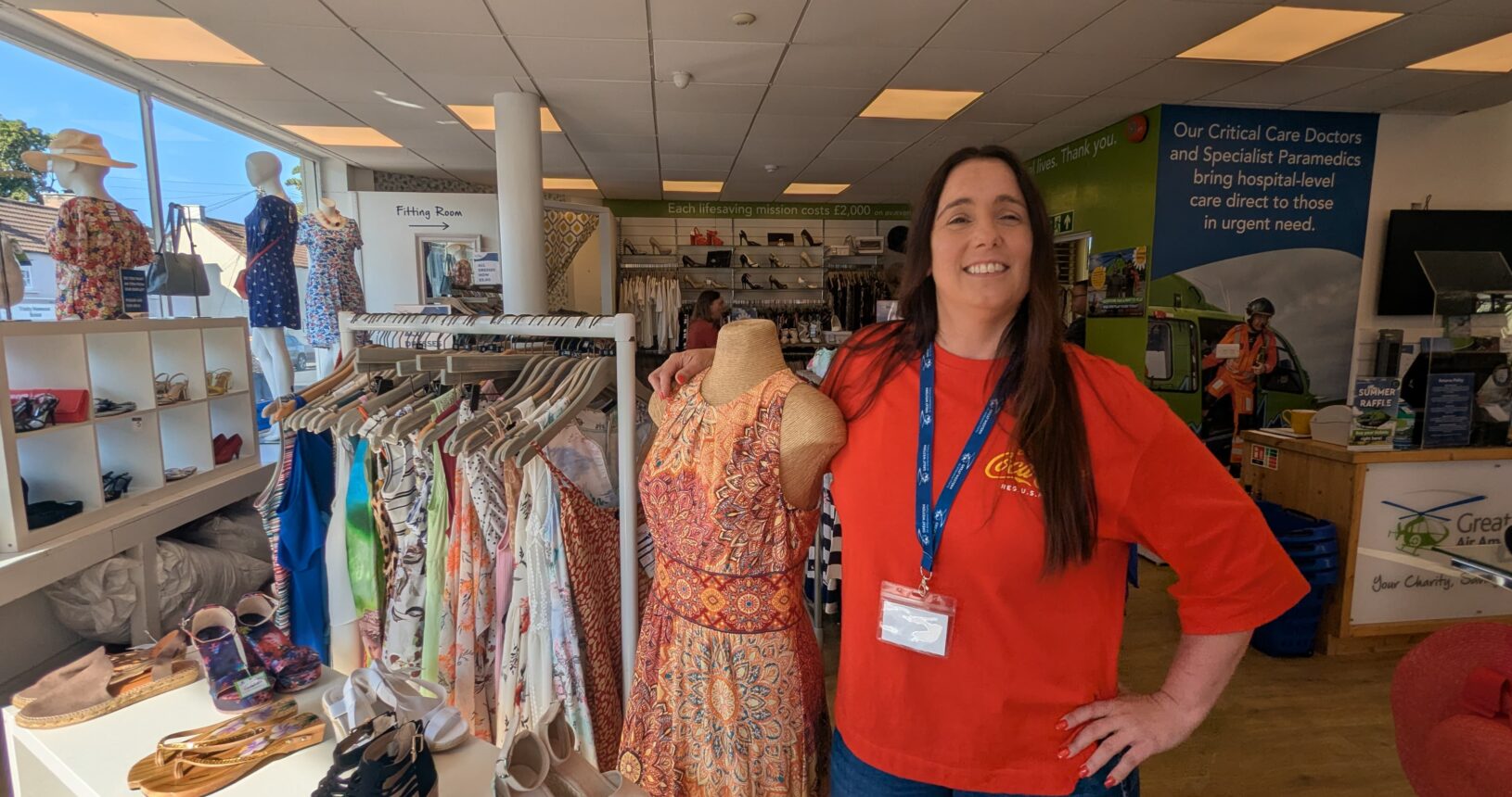 A corporate volunteer wearing a GWAAC lanyard stands proudly next to a dressed mannequin and clothing racks inside a bright Great Western Air Ambulance Charity shop.