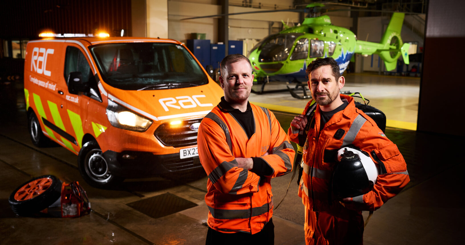 An RAC patrol worker standing side-by-side with a Great Western Air Ambulance Charity crew member inside an aircraft hangar, with a bright orange RAC breakdown van on the left and a green GWAAC helicopter in the background.