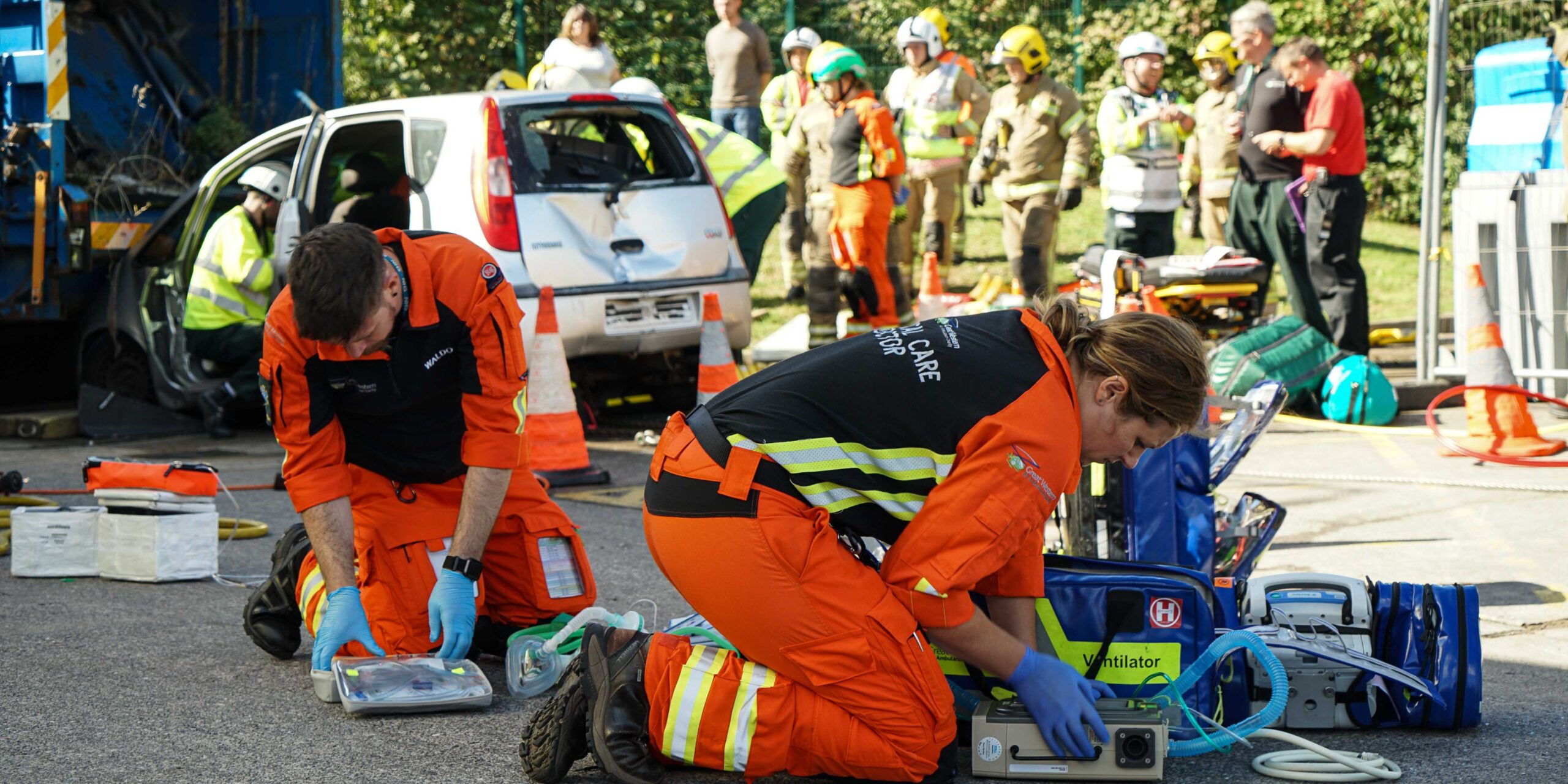 Two Great Western Air Ambulance Charity crew members on their knees working with pieces of equipment as they prepare to treat their patient. In the background a crushed car can be seen with lots of emergency service staff looking on as they work to get the patient from the car.