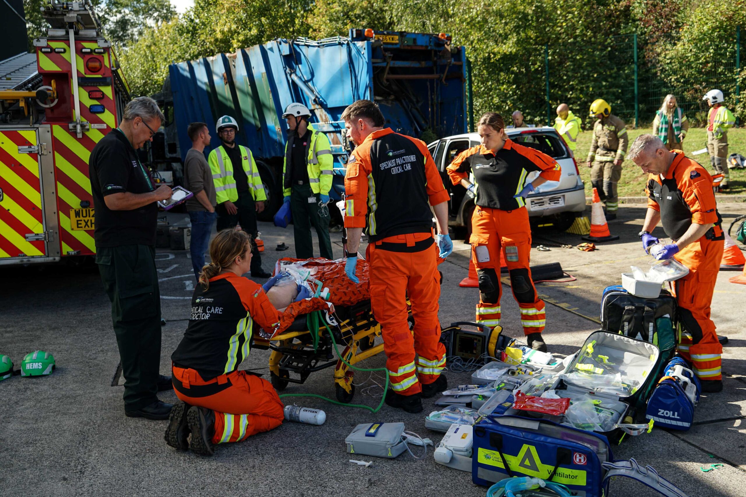 Great Western Air Ambulance Charity crew treat an injured patient on a stretcher. Medical kit is spread across the floor and four members of the team are treating their patient whilst an assessor overlooks and other emergency crews look on. They are stood next to the crashed lorry and car, as well as a fire engine.