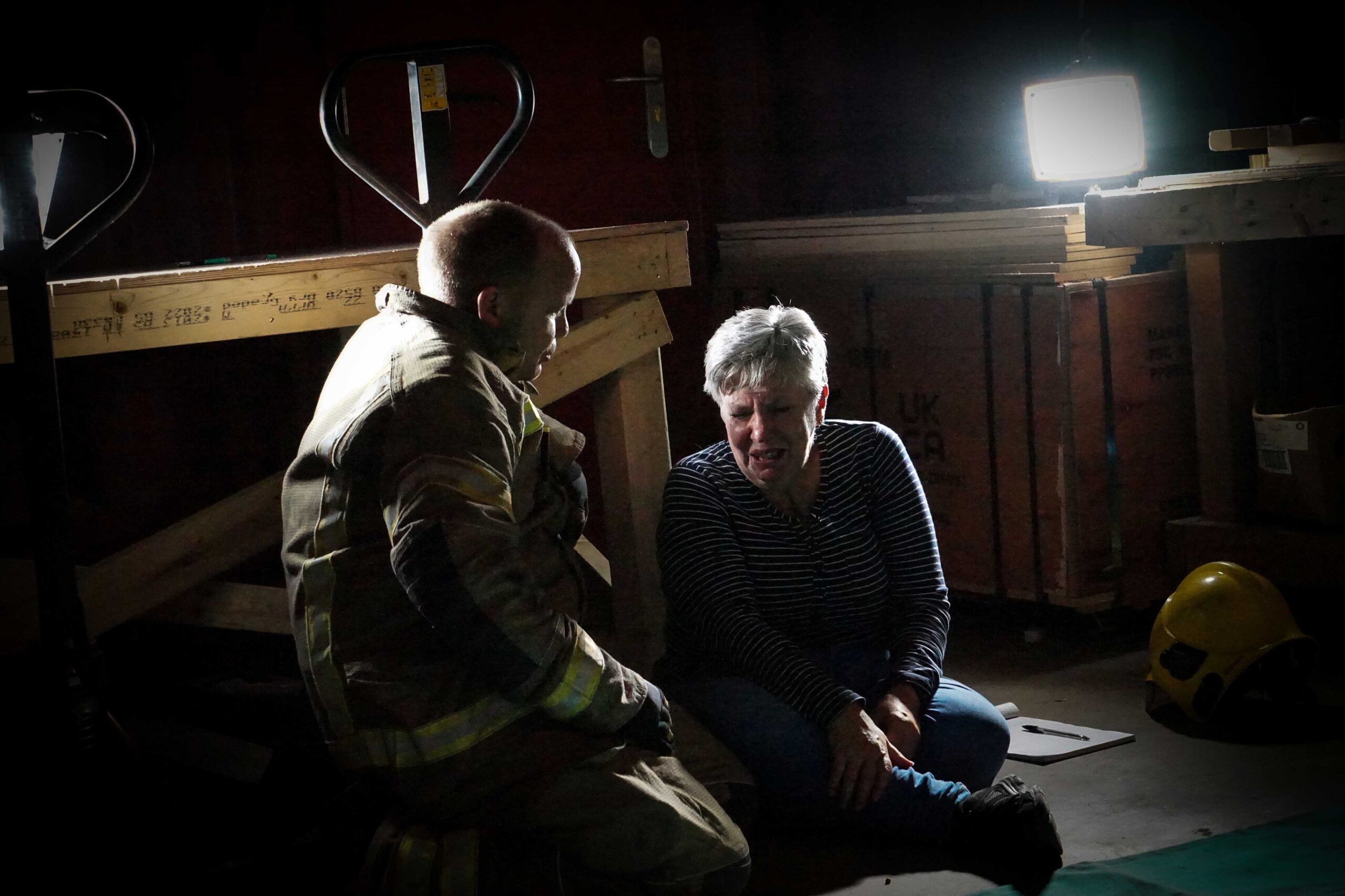 The bystander who found the patient is sat on the floor crying. She is being consoled by a firefighter as a spotlight lights the room.