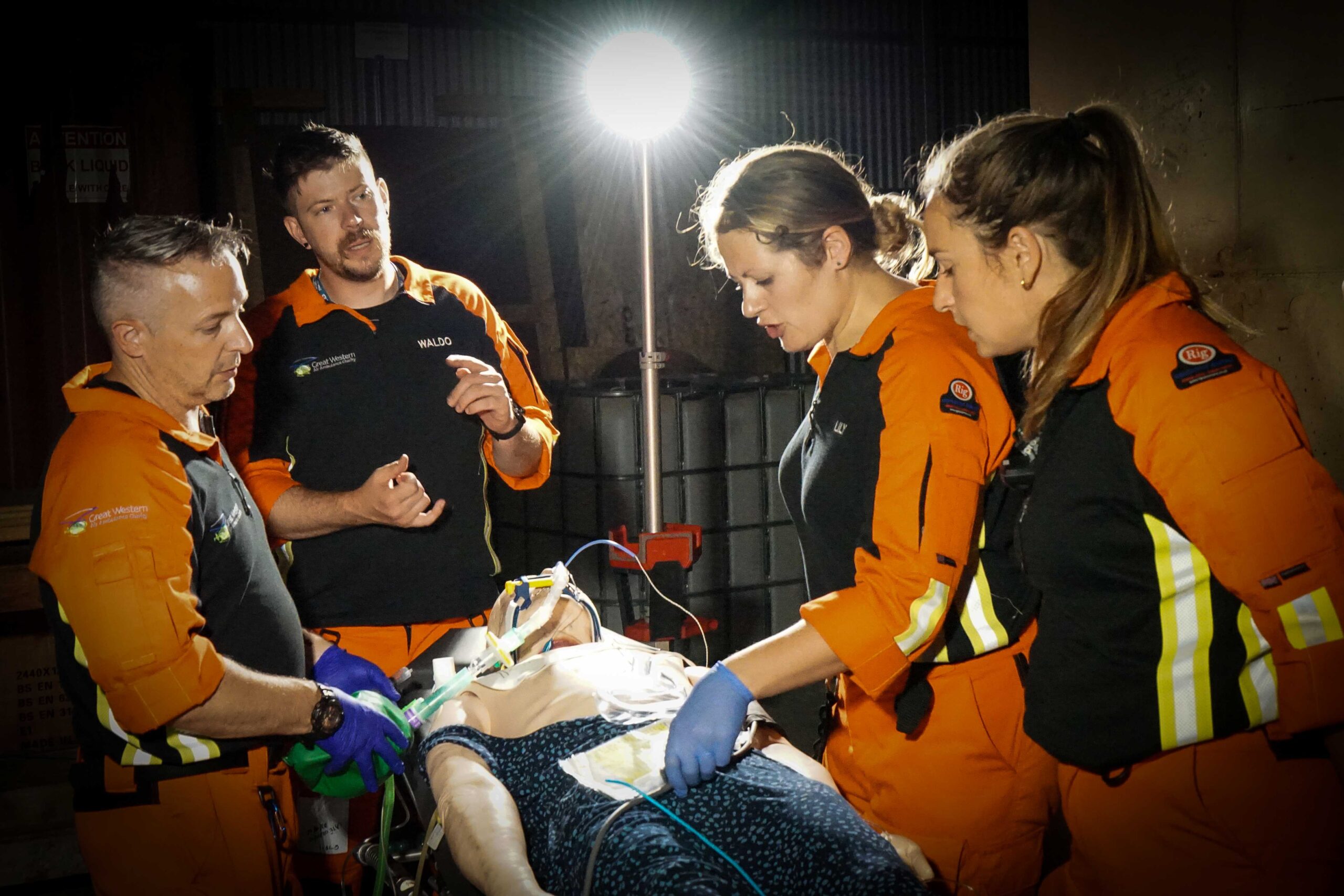 Great Western Air Ambulance Charity crew members treat their patient under a spotlight in a dark room. The team are mid conversation as they gather round a stretcher.