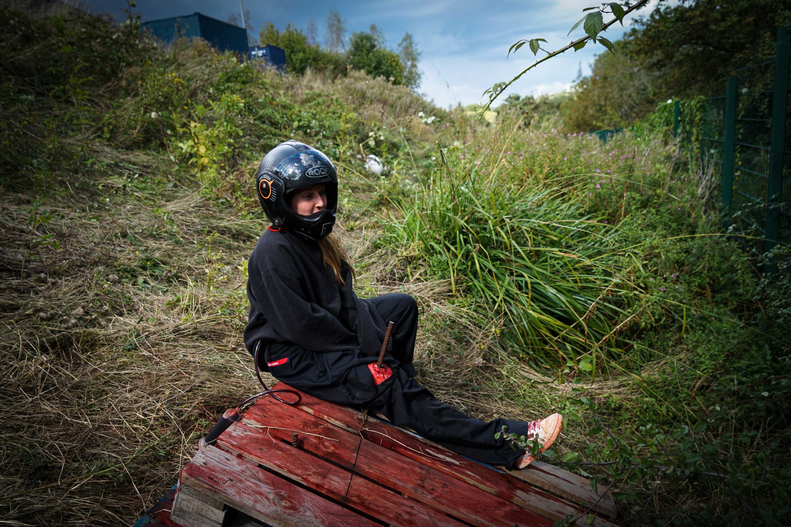 An injured motorcyclist is at the bottom of a ditch. They are still wearing their helmet whilst an iron rod is coming out of their leg. The wound is visible and fake blood can be seen on the wooden pallet next to them.