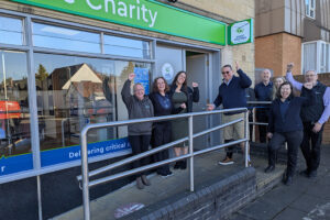 A group of people stand outside the newly opened Great Western Air Ambulance Charity shop in Brockworth, celebrating the occasion. The shop has a bright green sign with the charity's logo above the entrance. A man in sunglasses and a prosthetic leg holds a pair of scissors, having just cut the ribbon to officially open the store.