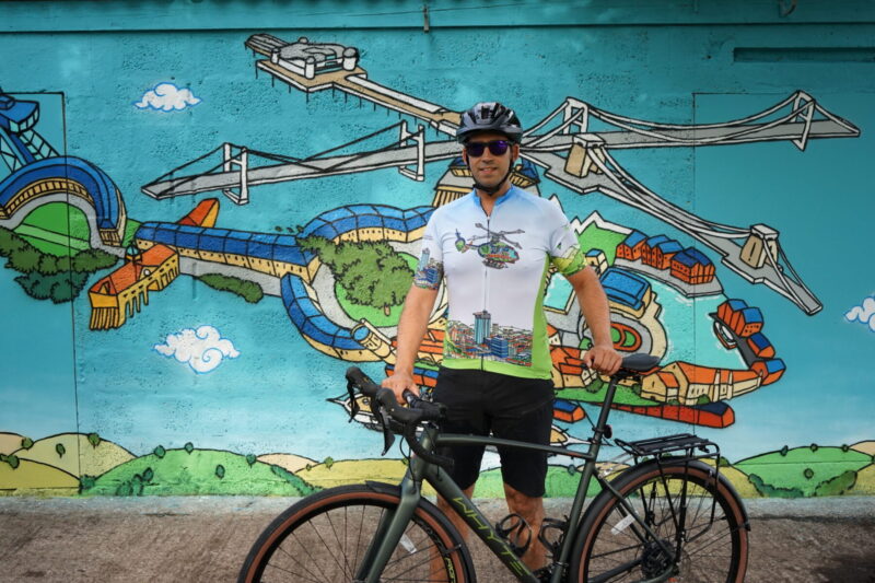 SPCC Matt Robinson wearing the GWAAC/ Presca/ Andy Council cycle jersey in front of the Andy Council helicopter mural