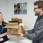 A smiling staff member from Great Western Air Ambulance Charity receives a box of donated picture frames from a cheerful supporter at a donation point indoors.
