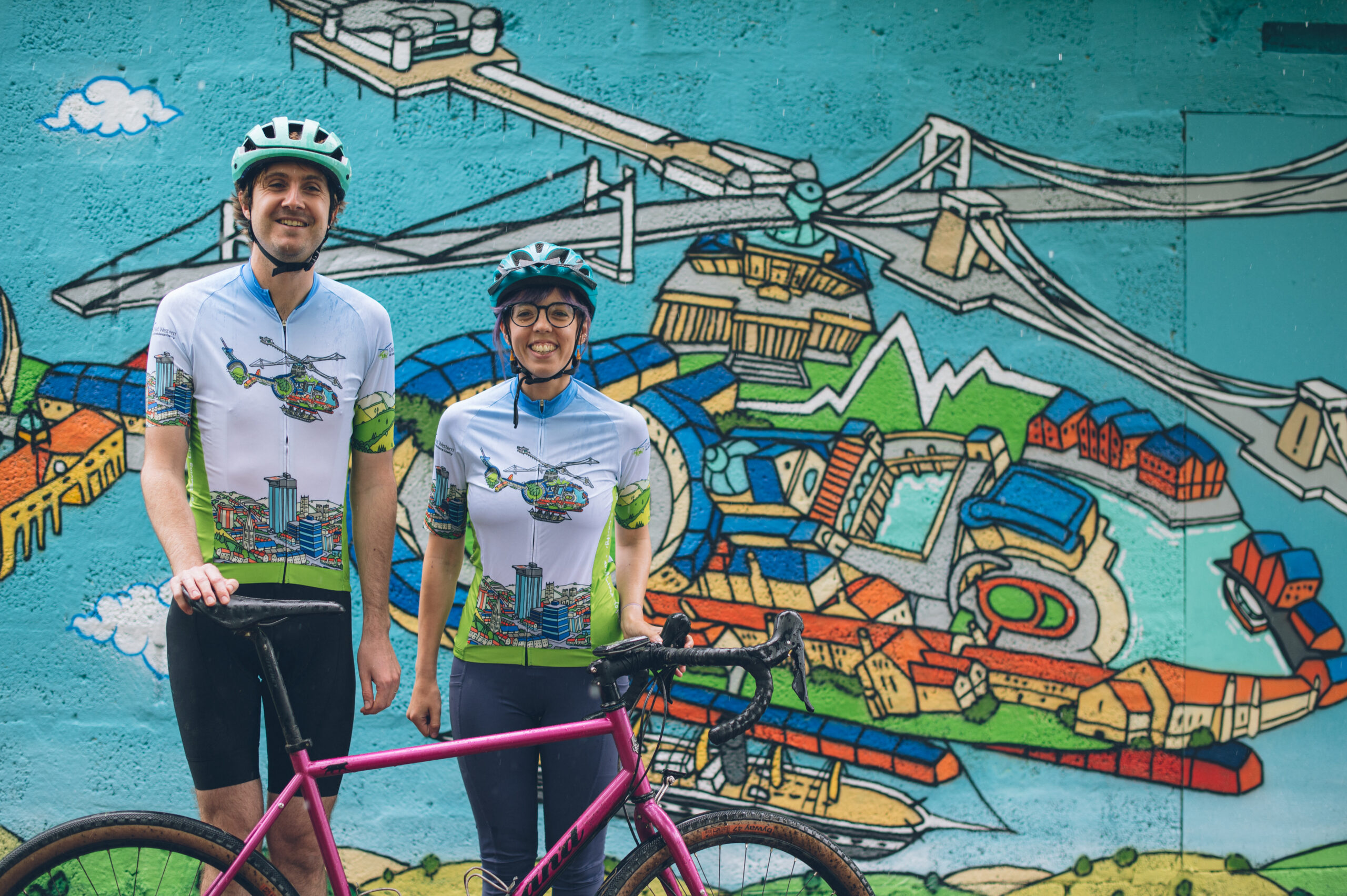 Man and lady cyclist in GWAAC/ Andy Council/ Presca cycle jersey in front of Andy Council mural at the GWAAC airbase