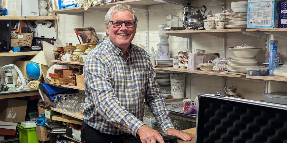 Volunteer Steve smiling while carrying out PAT testing on a donated electrical item in the back room of a Great Western Air Ambulance Charity shop, surrounded by shelves of donated goods and household items.