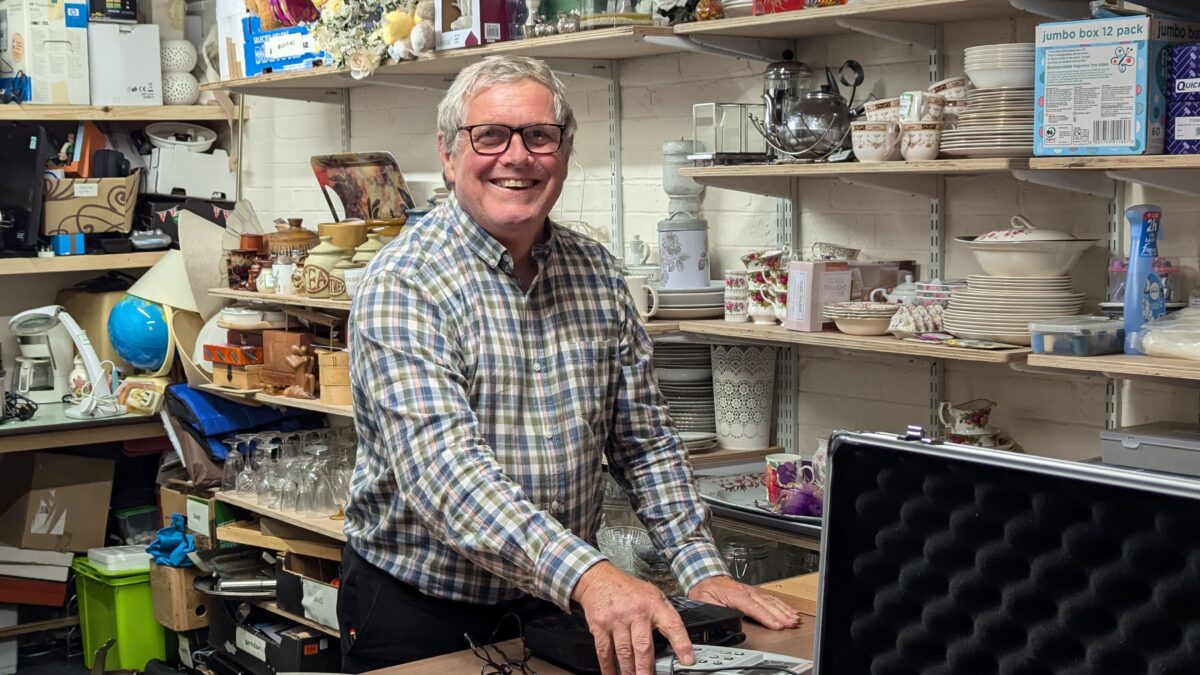Volunteer Steve smiling while carrying out PAT testing on a donated electrical item in the back room of a Great Western Air Ambulance Charity shop, surrounded by shelves of donated goods and household items.