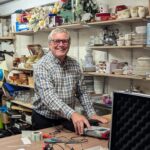 Volunteer Steve smiling while carrying out PAT testing on a donated electrical item in the back room of a Great Western Air Ambulance Charity shop, surrounded by shelves of donated goods and household items.