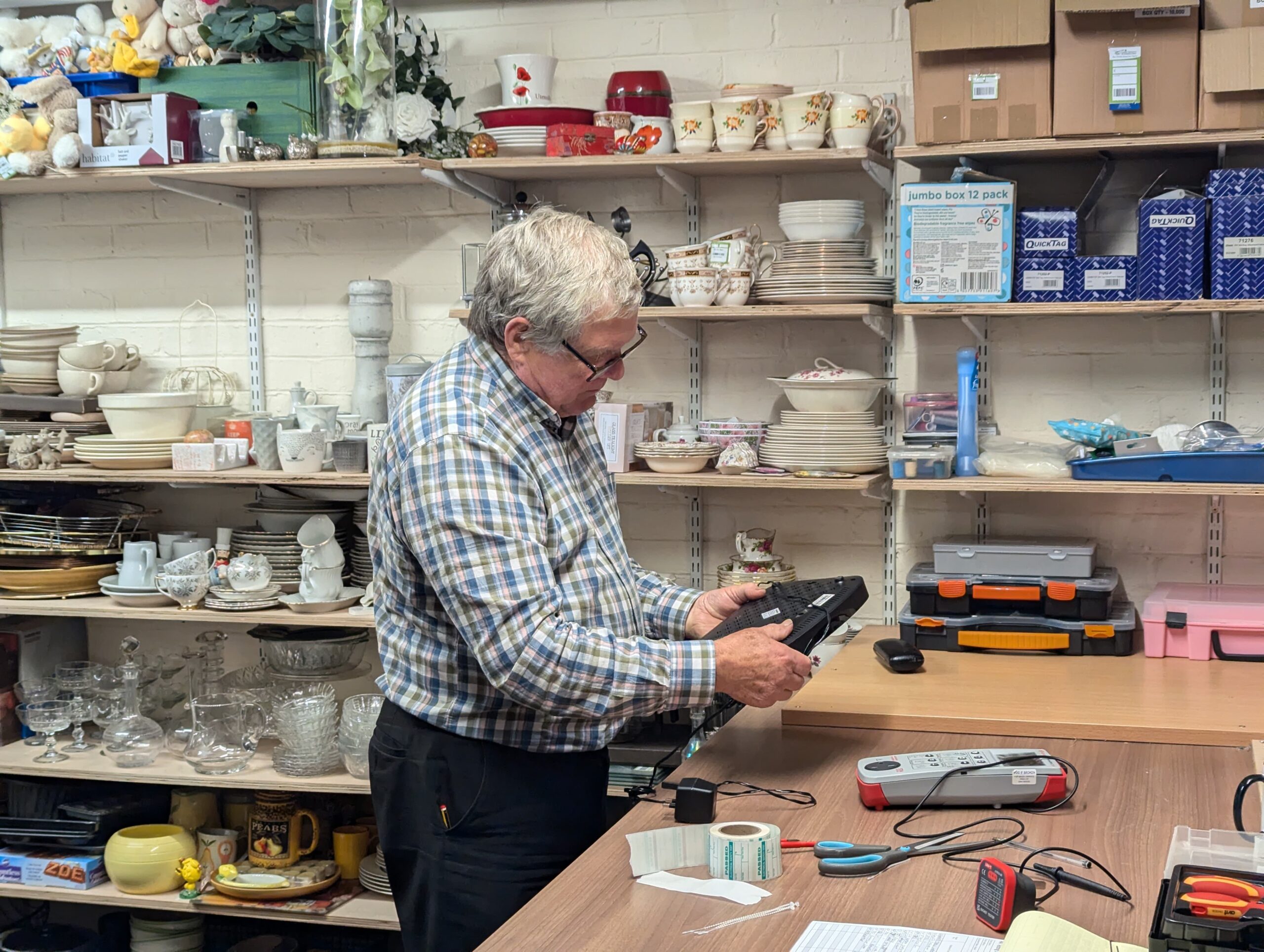 Volunteer Steve in the back room of a Great Western Air Ambulance Charity shop, carefully inspecting a donated electrical item while carrying out PAT testing. Shelves of donated kitchenware and household items are visible in the background.