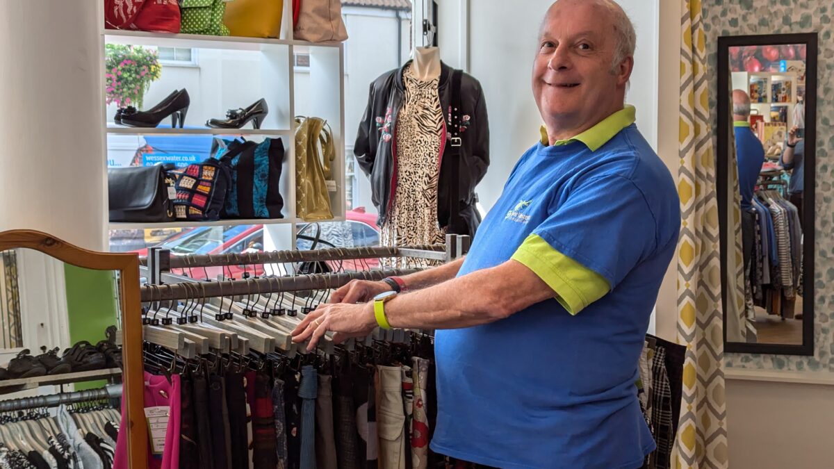 Volunteer Andy, wearing a blue and green Great Western Air Ambulance Charity t-shirt, smiles while organising a clothes rack in the Westbury-on-Trym shop. Behind him, handbags, shoes, and clothing are neatly displayed, with natural light streaming in through the window.