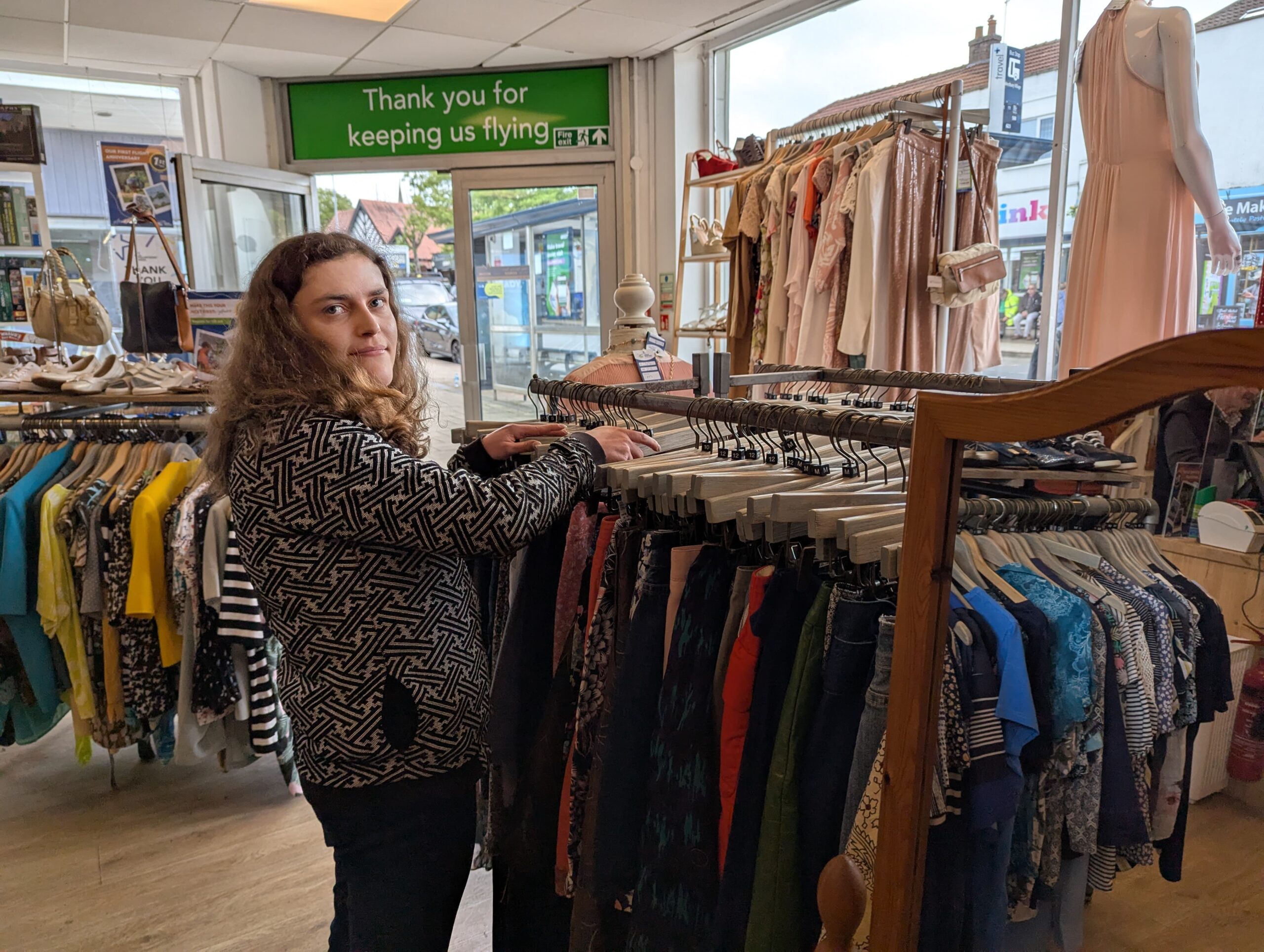 Retail volunteer, Joanna, inside Westbury-on-Trym's GWAAC shop, arranging clothes on a rail. The shop is bright, with racks of colourful clothing, shoes, and accessories visible in the background. A green sign above the doorway reads "Thank you for keeping us flying"