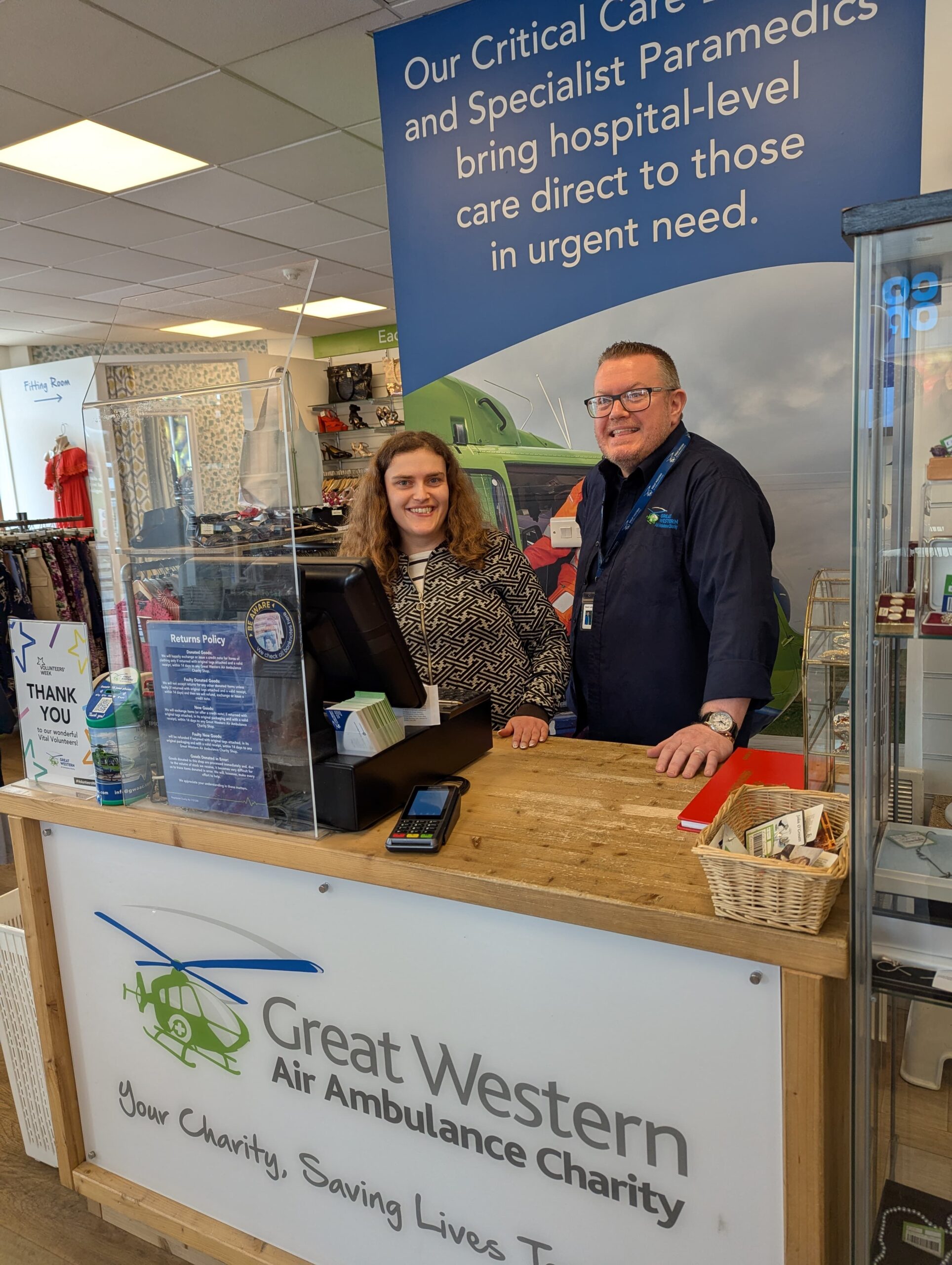Retail volunteer Joanna is standing behind the shop counter next to Shop Manager Stewart. Both are positioned in front of the till area. The counter has a card reader, a basket with items, and a returns policy sign. Clothing racks and other shop displays are visible in the background.