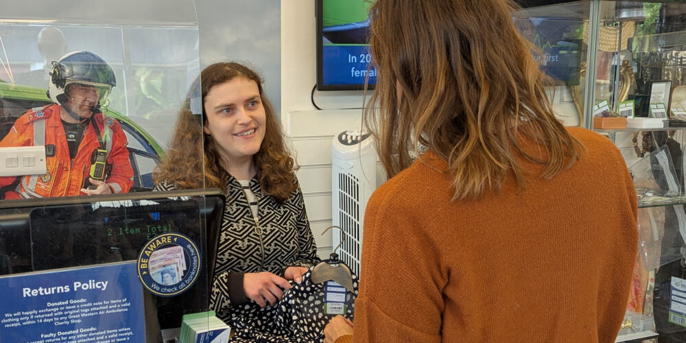 Retail volunteer Joanna is standing behind the shop counter, assisting a customer who is making a payment. The counter has a clear protective screen, a card reader, and a sign displaying the returns policy. Behind Joanna, there is a large poster featuring an air ambulance and text about providing hospital-level care in urgent situations. Shelves with merchandise and a TV screen are visible in the background.