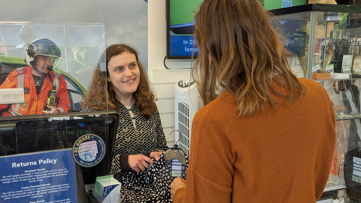 Retail volunteer Joanna is standing behind the shop counter, assisting a customer who is making a payment. The counter has a clear protective screen, a card reader, and a sign displaying the returns policy. Behind Joanna, there is a large poster featuring an air ambulance and text about providing hospital-level care in urgent situations. Shelves with merchandise and a TV screen are visible in the background.