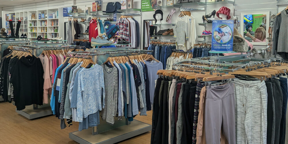 Interior of the recently refitted Great Western Air Ambulance Charity shop in Yate. The store features neatly arranged clothing racks with tops, trousers, and jackets in various colors and styles. Shelving along the back wall displays books, handbags, shoes, and accessories. The space has bright overhead lighting, a clean white ceiling, and light wood flooring, creating an open and organized layout. A few customers are visible browsing near the back.