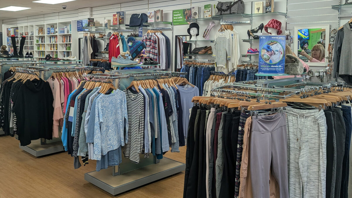 Interior of the recently refitted Great Western Air Ambulance Charity shop in Yate. The store features neatly arranged clothing racks with tops, trousers, and jackets in various colors and styles. Shelving along the back wall displays books, handbags, shoes, and accessories. The space has bright overhead lighting, a clean white ceiling, and light wood flooring, creating an open and organized layout. A few customers are visible browsing near the back.