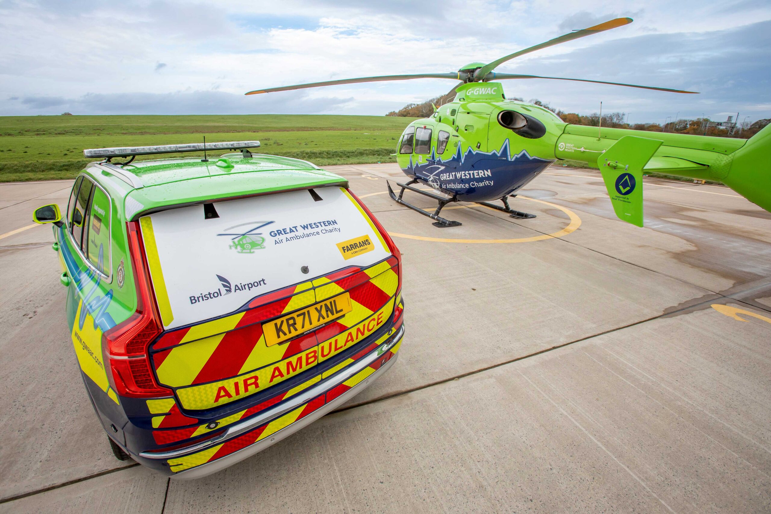 The back of Great Western Air Ambulance's new Volvo XC90 critical care car. The rear windshield is white and has the logos of Great Western Air Ambulance Charity, Bristol Airport and Farrans on it. Under the license plate, the words 'air ambulance' are there to alert people that the car is an emergency vehicle. The car is parked alongside the green and blue helicopter.