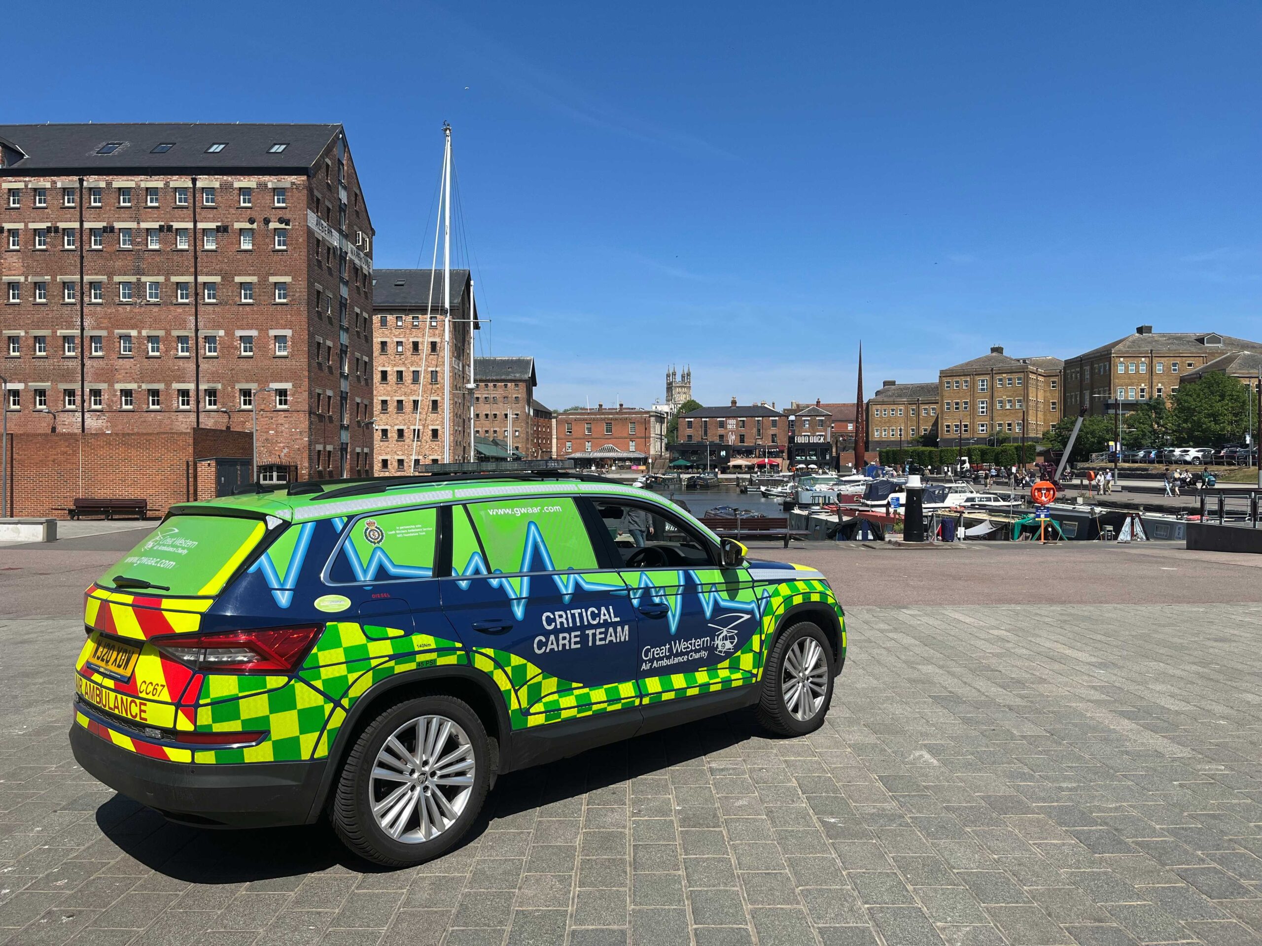 A green, blue and yellow critical care car pictured in front of Gloucester Docks, with boats and buildings in the background