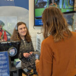 Retail volunteer Joanna is standing behind the shop counter, assisting a customer who is making a payment. The counter has a clear protective screen, a card reader, and a sign displaying the returns policy. Behind Joanna, there is a large poster featuring an air ambulance and text about providing hospital-level care in urgent situations. Shelves with merchandise and a TV screen are visible in the background.