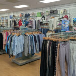 Interior of the recently refitted Great Western Air Ambulance Charity shop in Yate. The store features neatly arranged clothing racks with tops, trousers, and jackets in various colors and styles. Shelving along the back wall displays books, handbags, shoes, and accessories. The space has bright overhead lighting, a clean white ceiling, and light wood flooring, creating an open and organized layout. A few customers are visible browsing near the back.