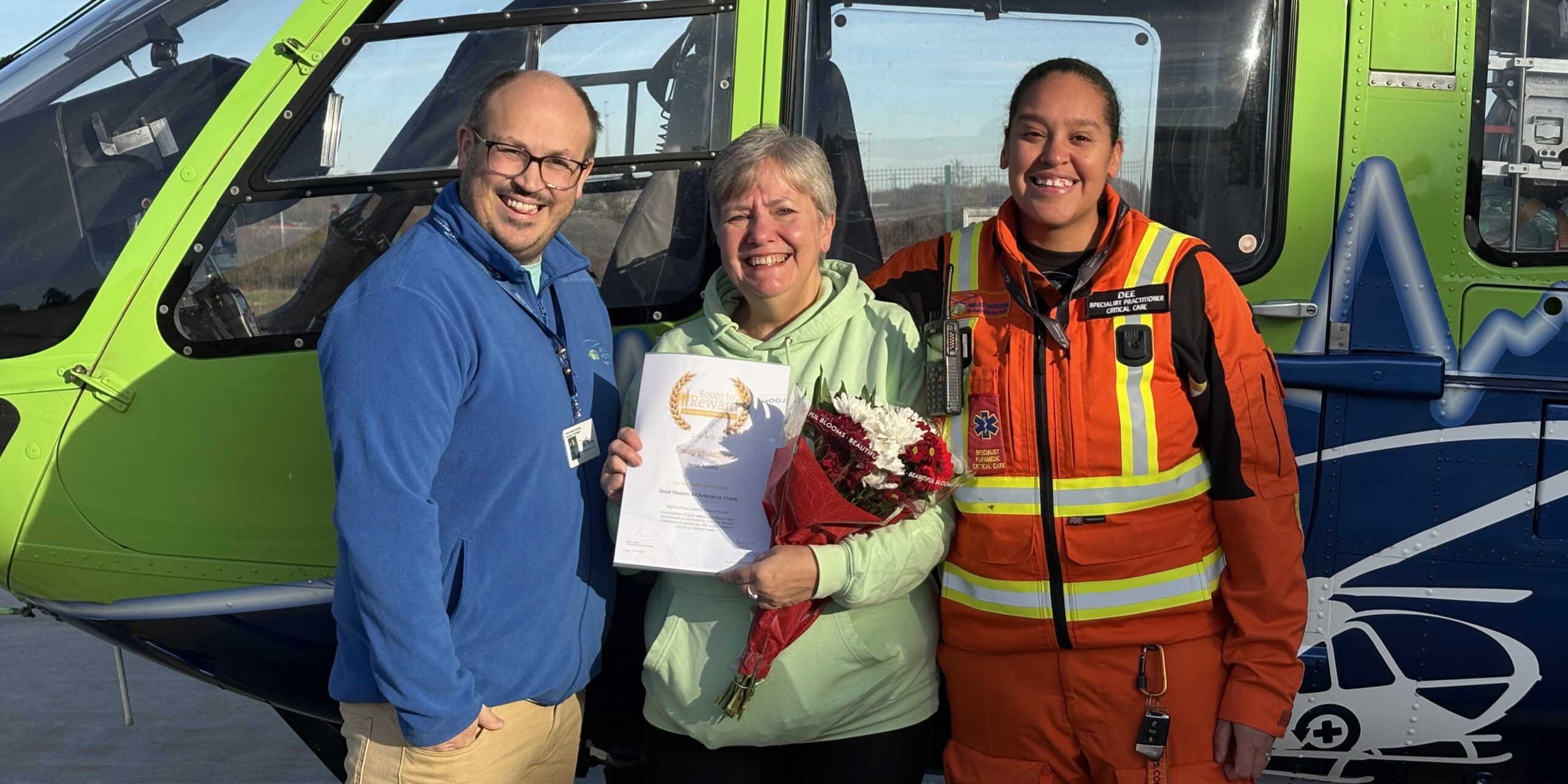 Janet poses with her certificate and flowers in front of the GWAAC green and blue helicopter, alongside Volunteer Coordinator Dan and Specialist Paramedic in Critical Care Dee