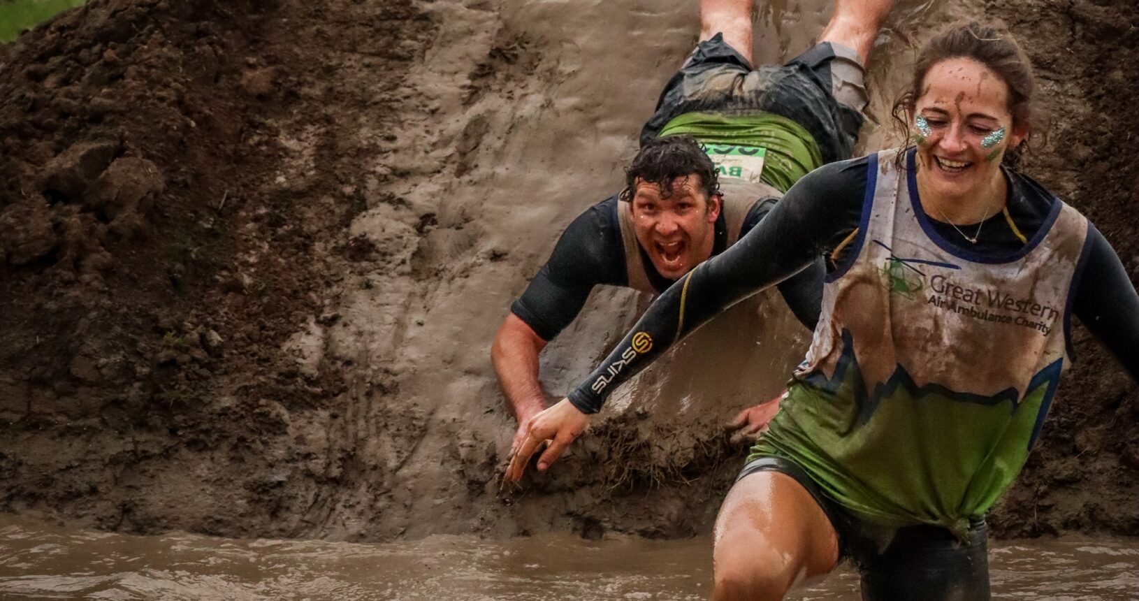 A man and a lady in GWAAC running vests negotiating a mud slide and mudbath at the Mud Master event