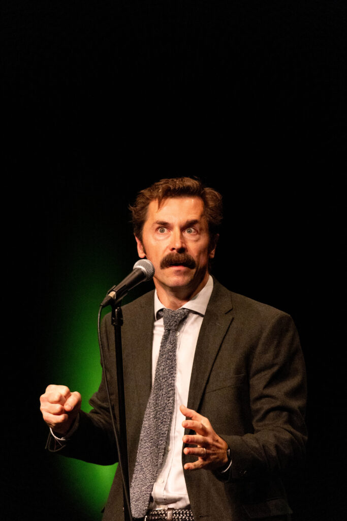 Mustached comedian Mike Wozniak preforming in a woolen suit and tie at Heli Laughs 2023 at The Old Vic Bristol